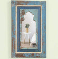 Distressed wooden mirror with a blue shade hanging on a wall with a golden planter showing in the mirror reflection