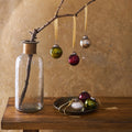 A glass vase with a branch coming from the top with 3 vintage glass baubles hanging from the branch, and 3 glass baubles underneath on a metal plate